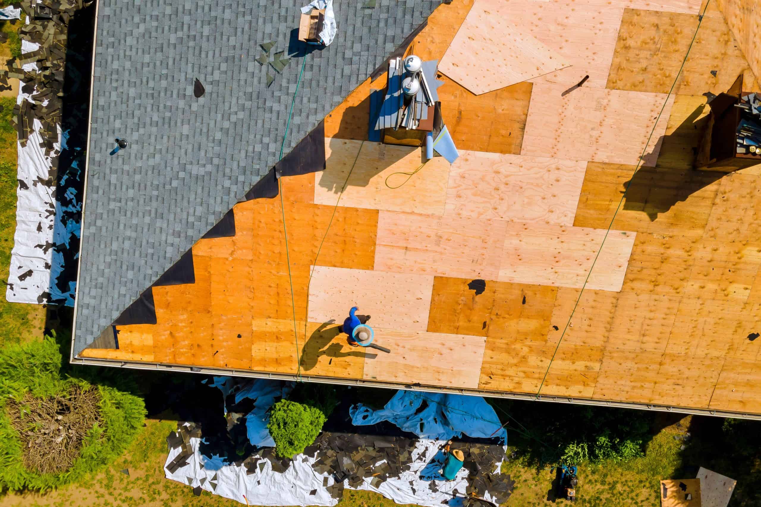 Aerial view of a roof under construction. Two workers in hard hats are installing plywood sheets. Part of the roof is covered with shingles. Tarps and debris are visible on the ground below.
