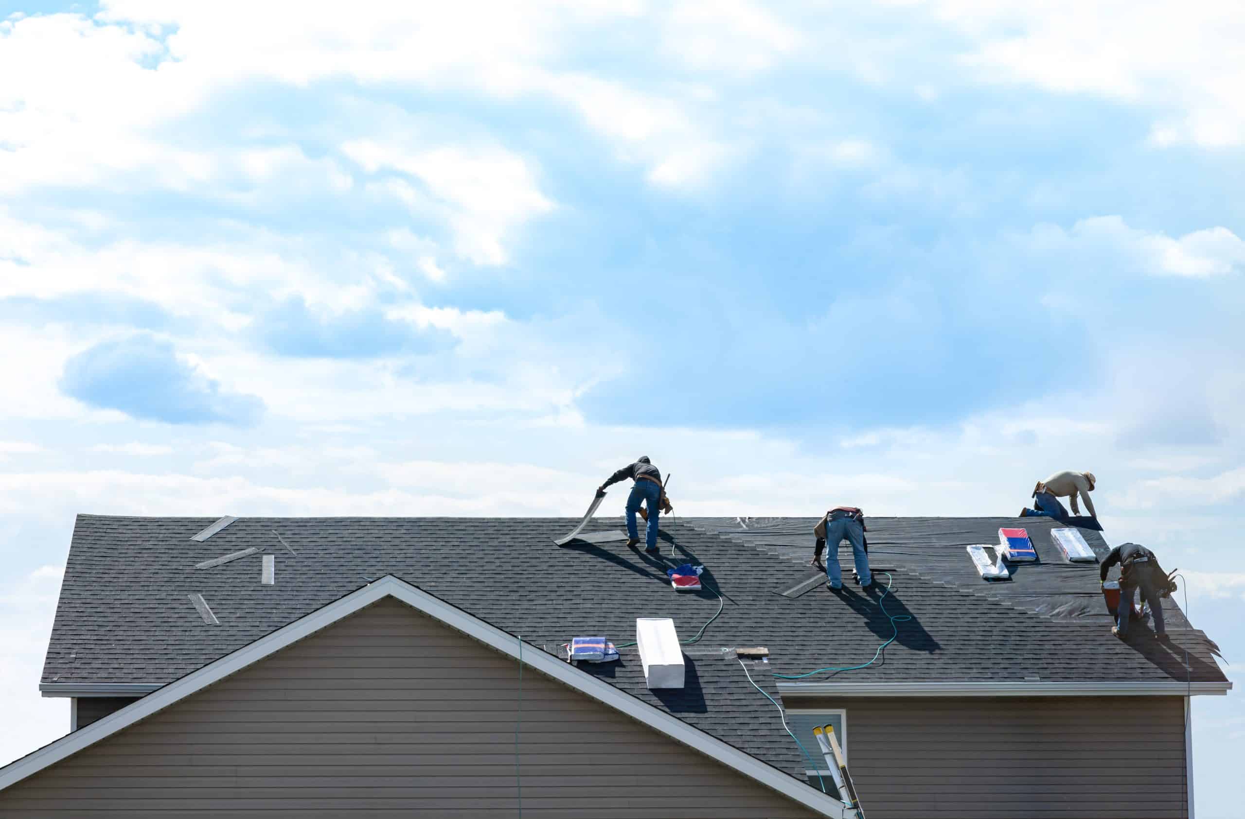 Roofers working on a two-story house, installing new shingles. Three workers are focused on the dark roof against a cloudy sky backdrop. Equipment and materials are scattered across the roof surface.